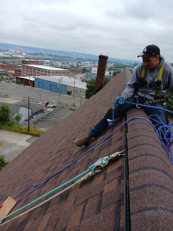 Roofer in a safety harness, tied off with ropes, working on a steep brown asphalt shingle roof