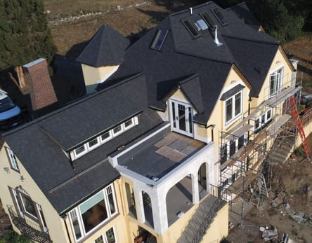 Aerial view of a large home whose roof combines steep asphalt shingle sections, dormers, skylights, a chimney, and a low-slope porch roof