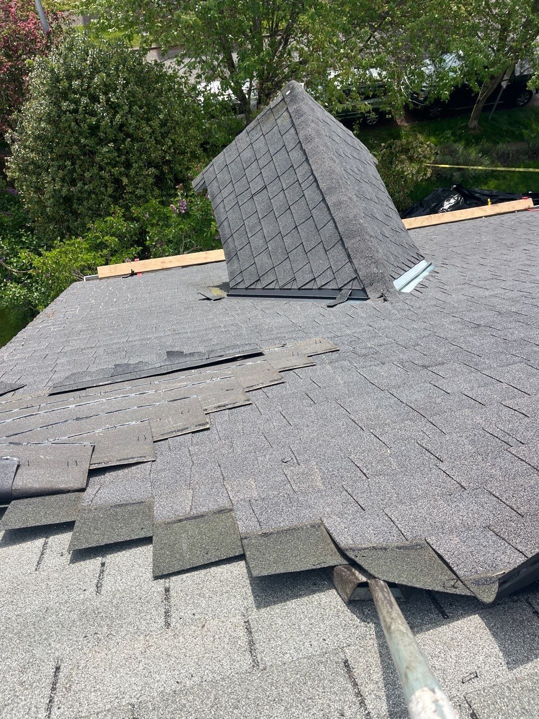 Old gray asphalt shingles being pried up in rows during a roof tear-off, with a dormer and trees in the background