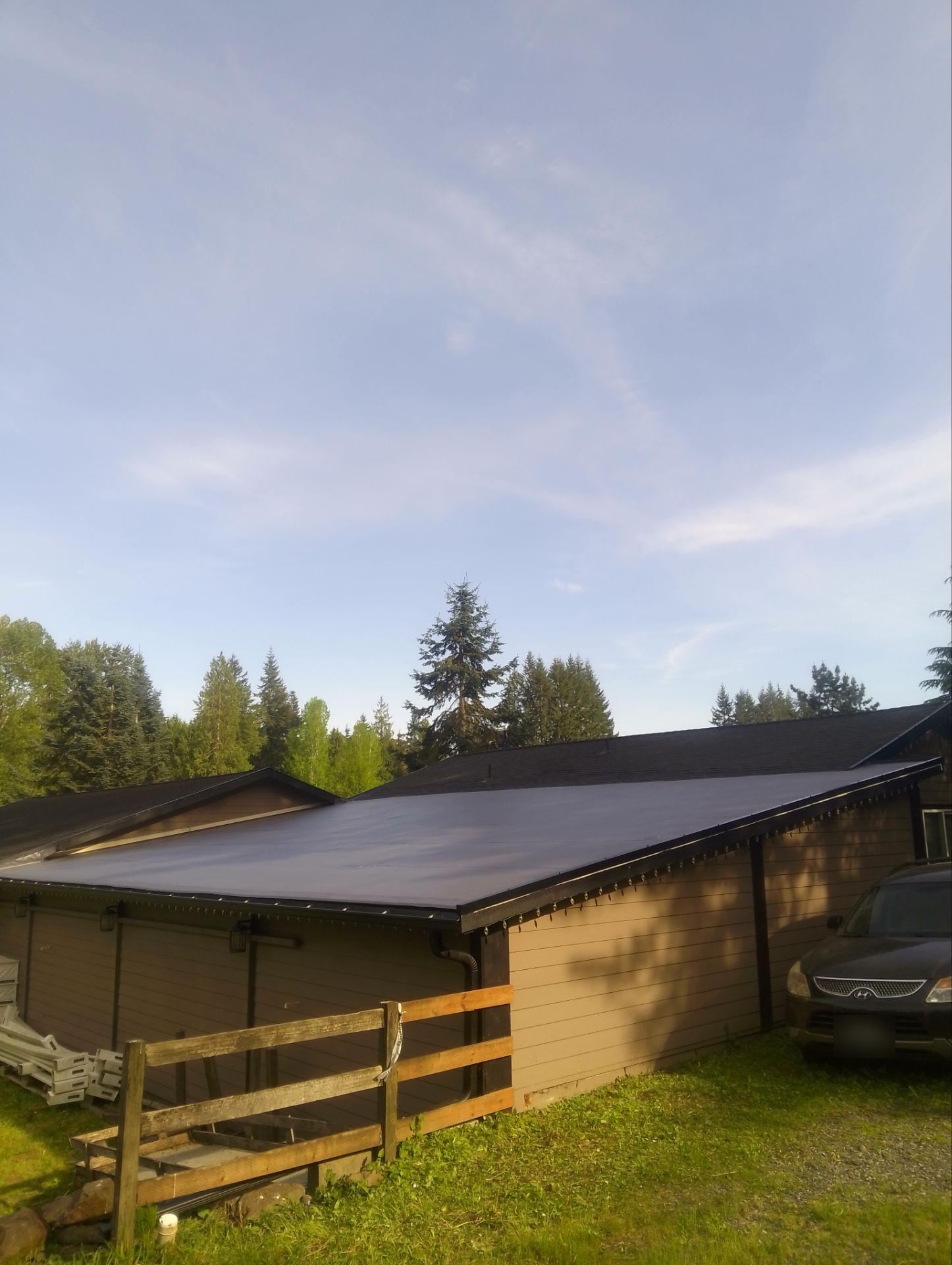 Dark low-slope roof on a long building with wood siding, a fence and tall evergreens